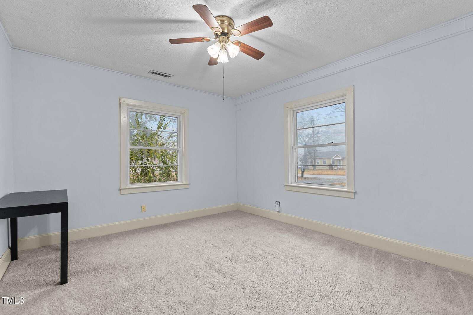An empty room with light blue walls and beige carpet. Two windows provide natural light, and a ceiling fan with wooden blades is centered on the ceiling. A small black table is in the corner near one window.