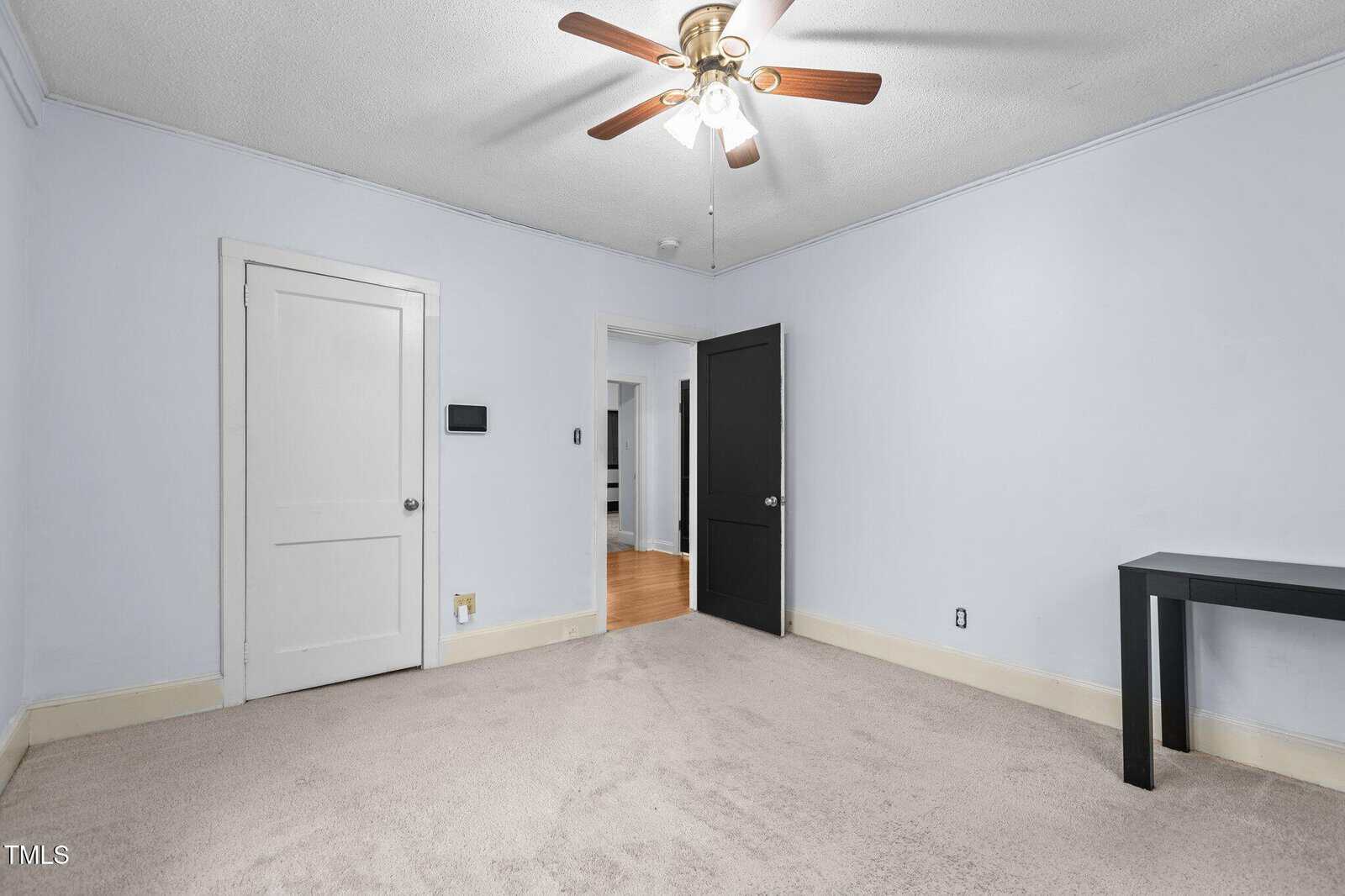 An empty room with light blue walls and beige carpet. A ceiling fan with wooden blades is centered above. The room has a white door and a black door, with a narrow black table against one wall. A section of hardwood floor is visible through the doorway.