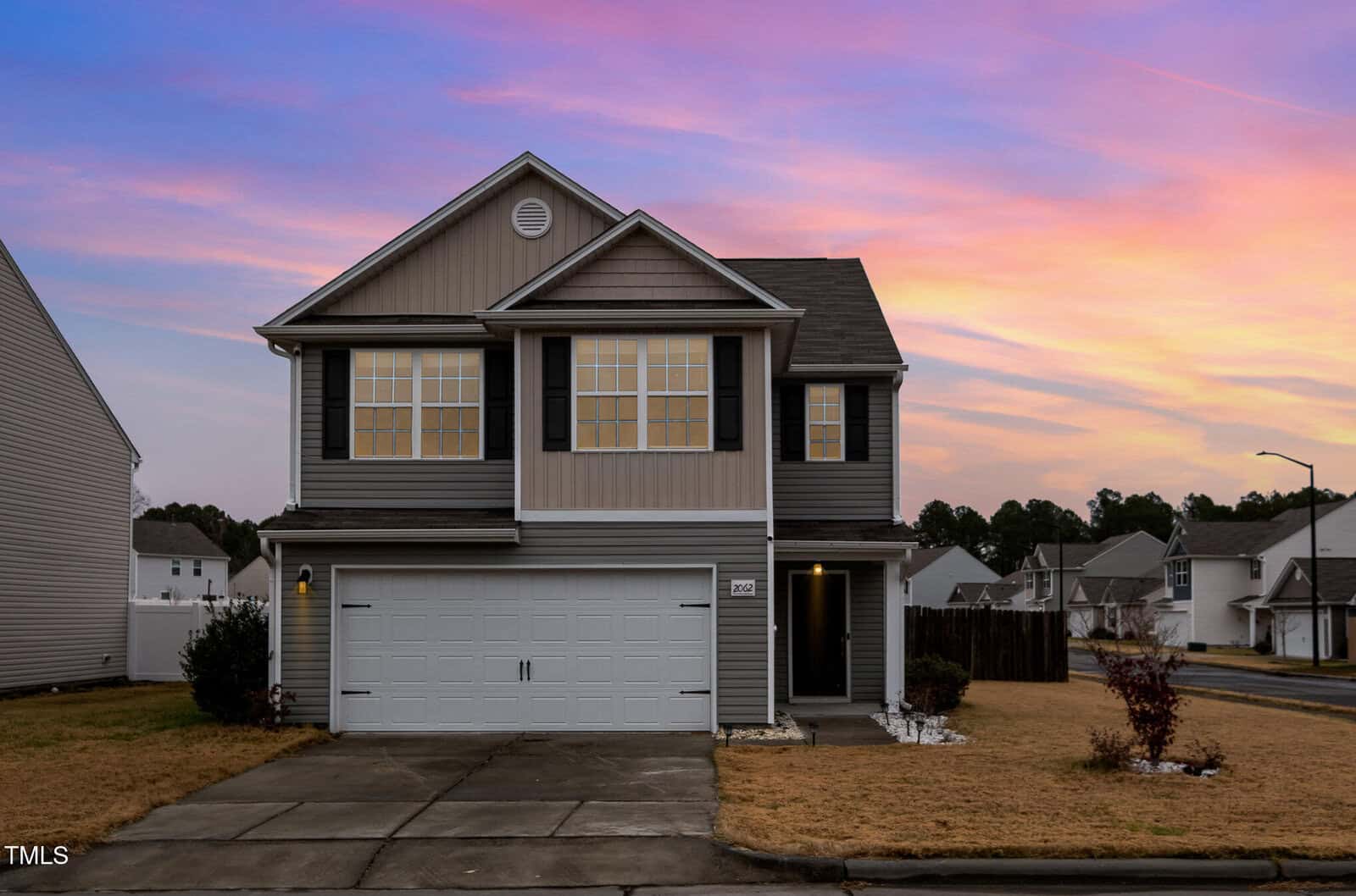 Two-story suburban house with a gray exterior and white trim, featuring a double garage and a small front garden. The sky is filled with a sunset palette of pink, orange, and purple hues. The house is situated in a residential neighborhood.