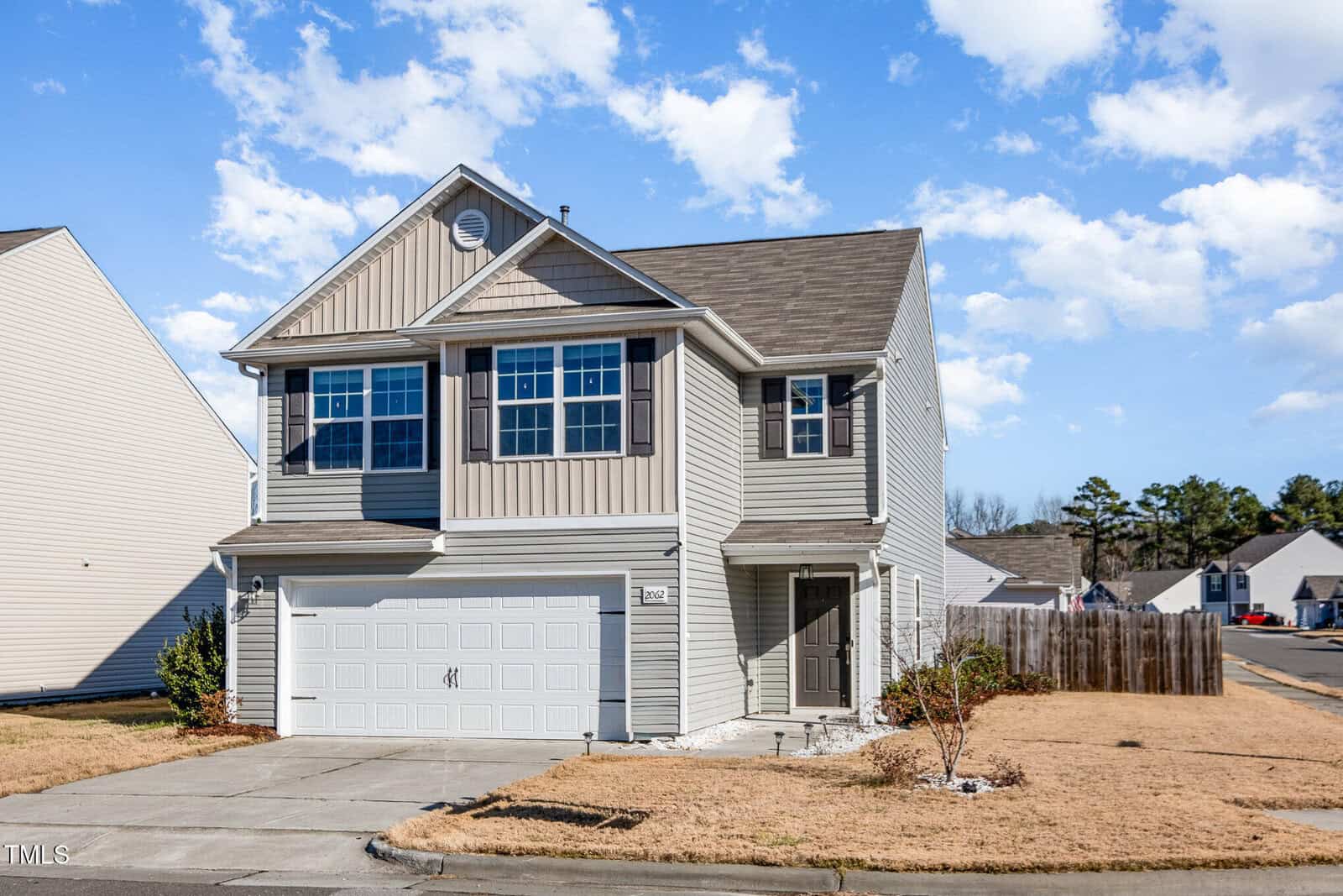 A two-story suburban house with a gray exterior and a two-car garage. The house features multiple windows and a small front porch. It is situated in a neighborhood with similar-style homes under a partly cloudy sky.