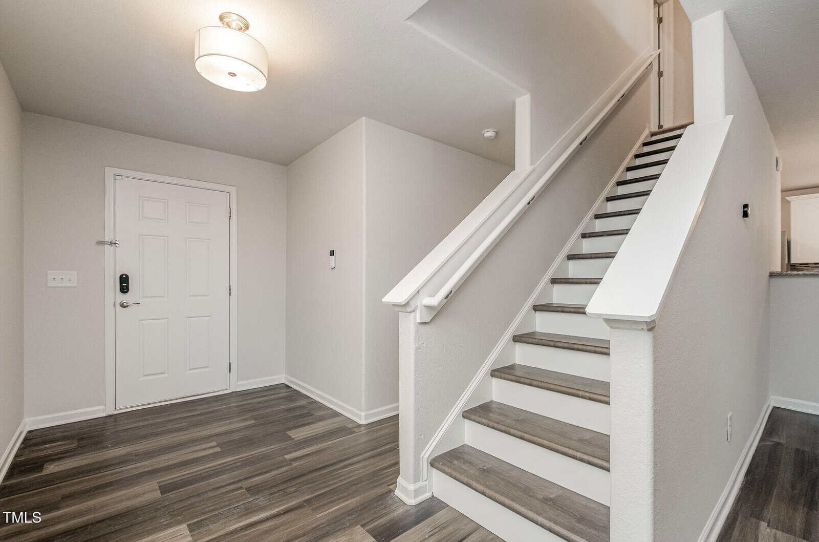 A modern hallway with light grey walls and dark wood flooring. There is a white door on the left and a staircase with white railings and dark steps on the right. A ceiling light fixture is visible above the entryway.