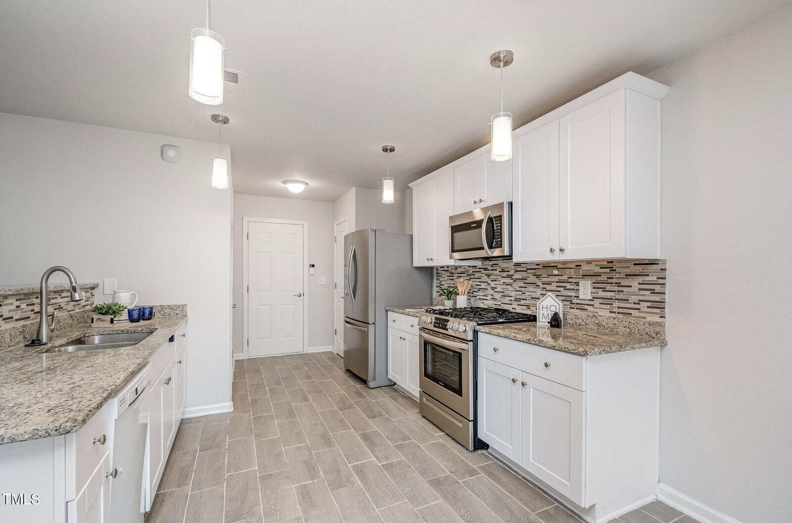 A modern kitchen with ceramic floor tiles, white cabinets, and granite countertops. Stainless steel appliances include a refrigerator, microwave, and oven. Pendant lights hang from the ceiling, and a multicolored tile backsplash adds contrast.