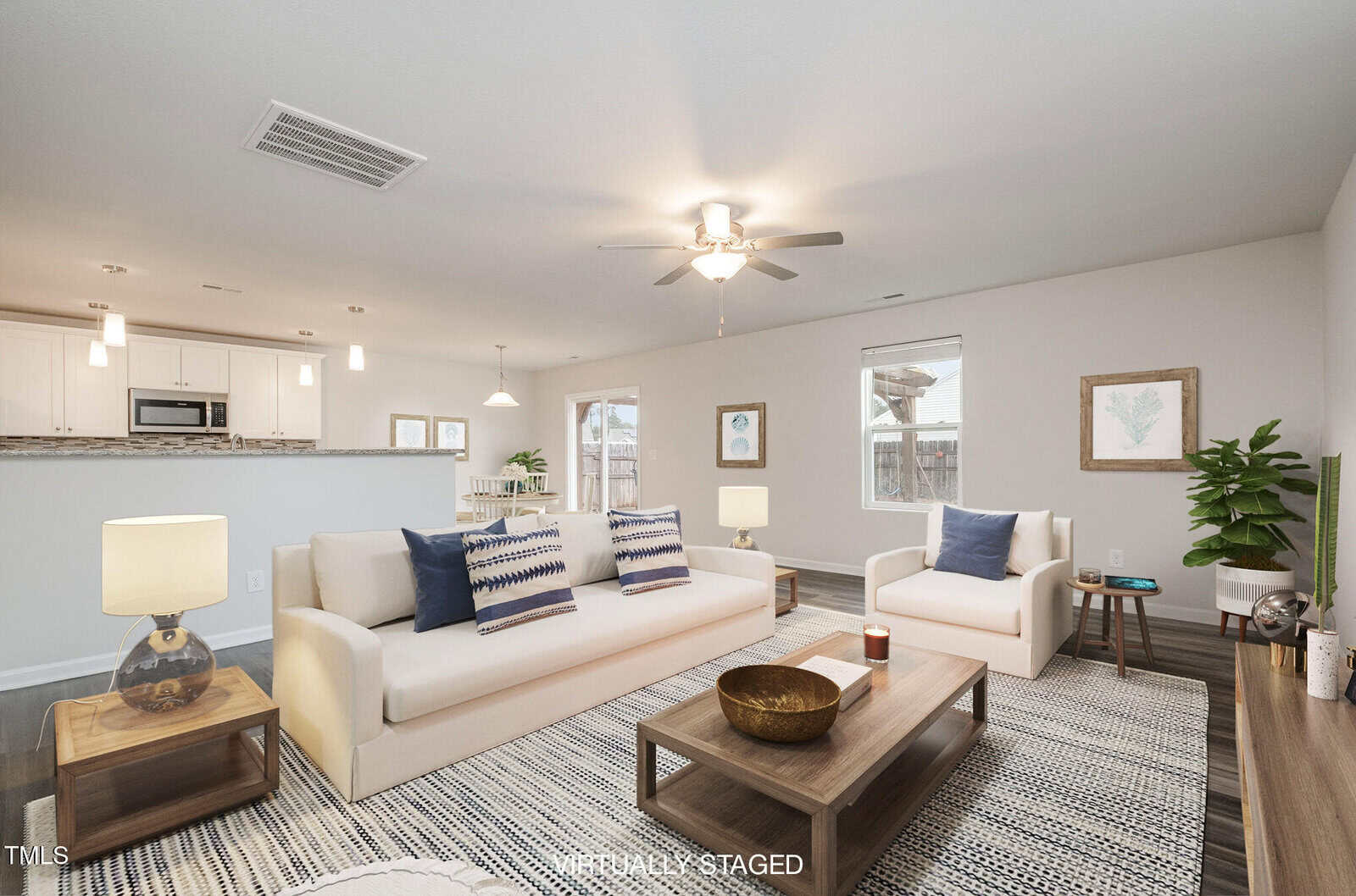 A modern living room features a white sofa and armchair with blue-patterned cushions. There are wooden coffee and side tables, a ceiling fan, and two framed artworks on the wall. A kitchen area is visible in the background. The room is virtually staged.
