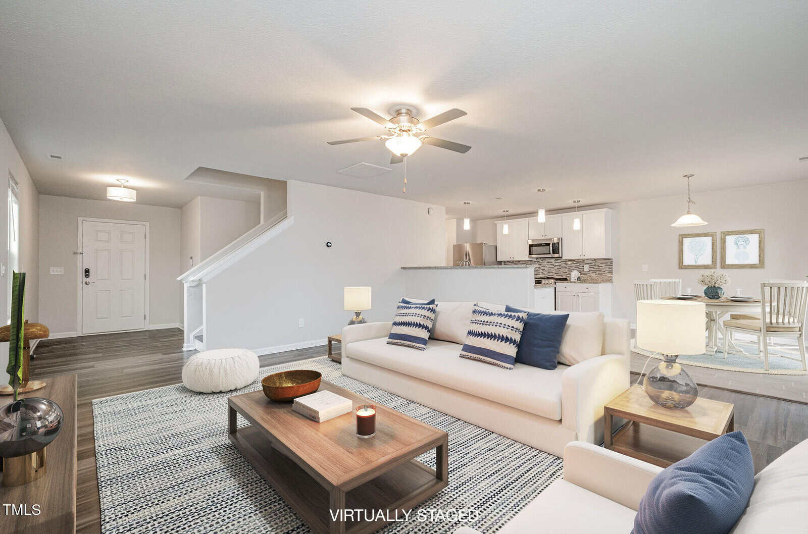 A modern, open-concept living area featuring a white sofa with patterned pillows, a wooden coffee table, and a rug. A ceiling fan is above. The space extends to a dining area and kitchen. A staircase is visible, and the room is well-lit with staged decor.