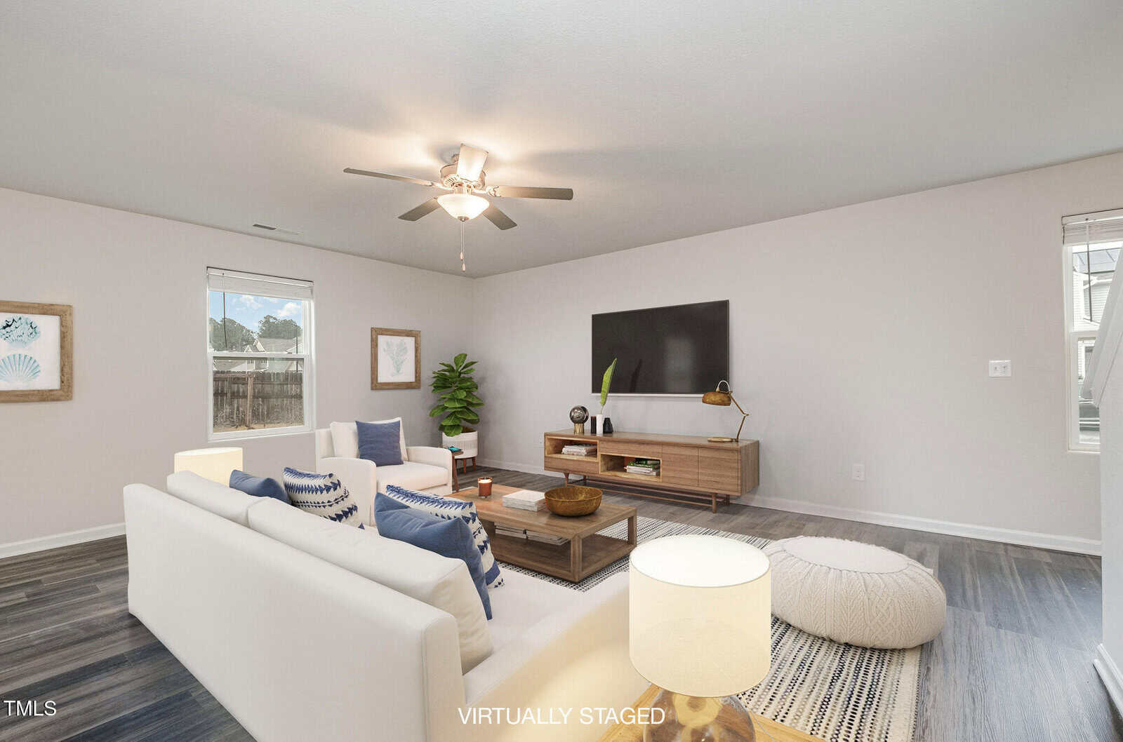 A virtually staged living room with a white sectional sofa, a wooden coffee table, and a large flat-screen TV on a wooden stand. There's a ceiling fan, a potted plant, a pouf, and a lamp on a side table. Art pieces adorn the walls.