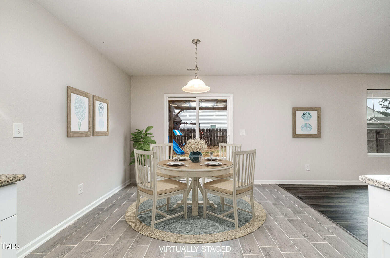Virtually staged dining room with a round table set for four. The room features light-colored walls, wood-look flooring, and framed artwork on the walls. Sliding glass door leads to an outdoor area with a view of a swing set and a fence.