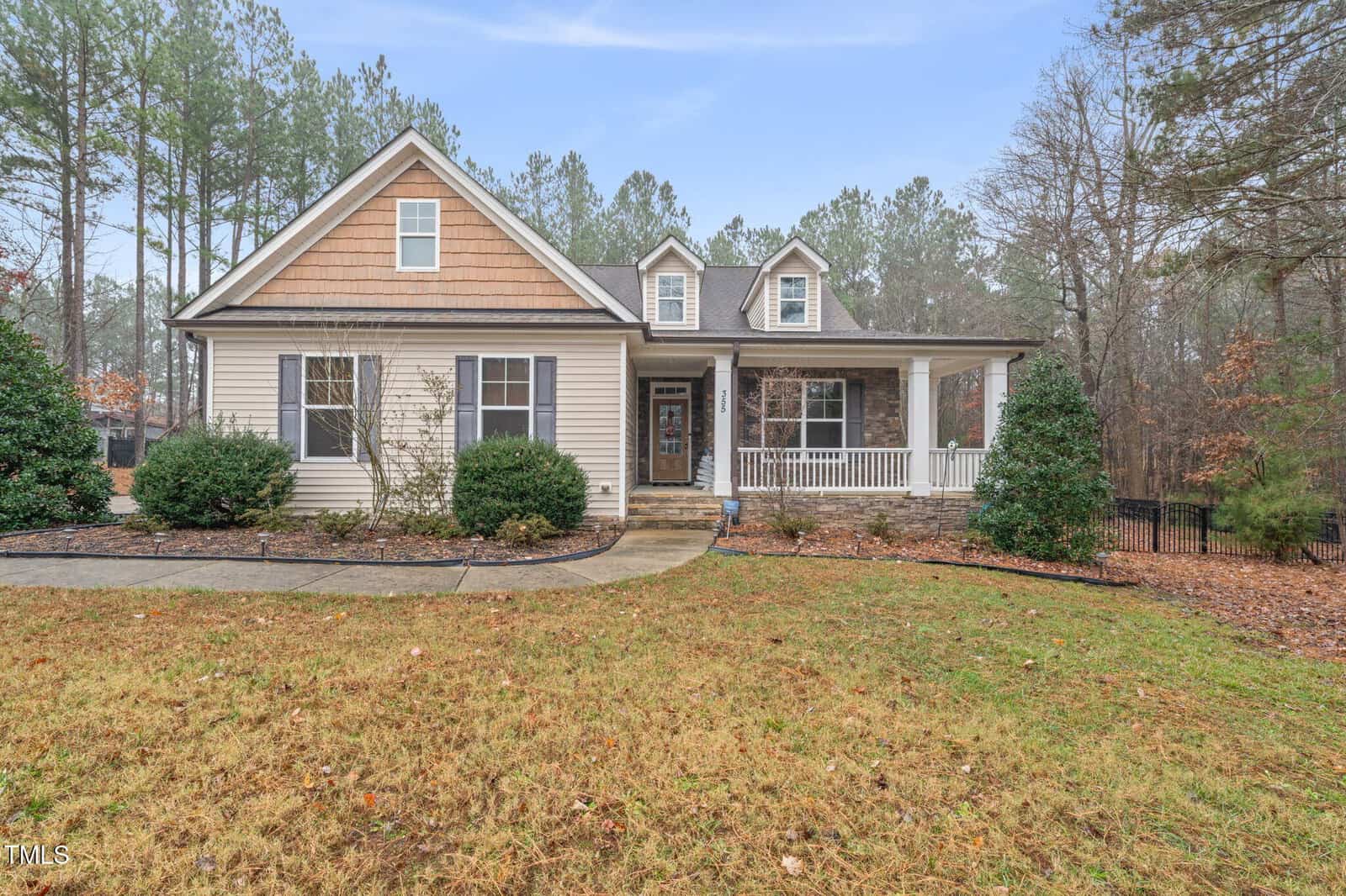 Single-story house with beige siding, stone accents, and a covered front porch, set on a grassy yard with shrubs and bordered by tall pine trees. A curved walkway leads to the front door.