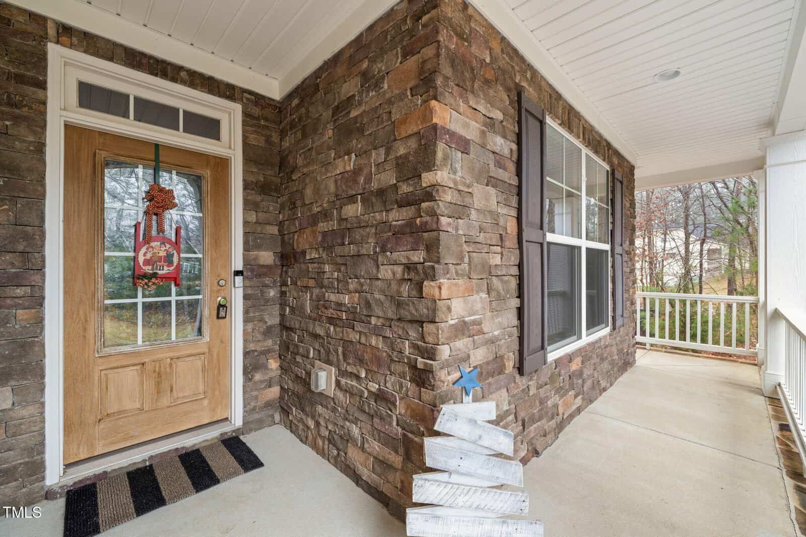 A covered front porch with stone walls, a wooden front door decorated with a holiday wreath, a large window with shutters, a doormat, and a small white wooden tree decoration on the concrete floor.