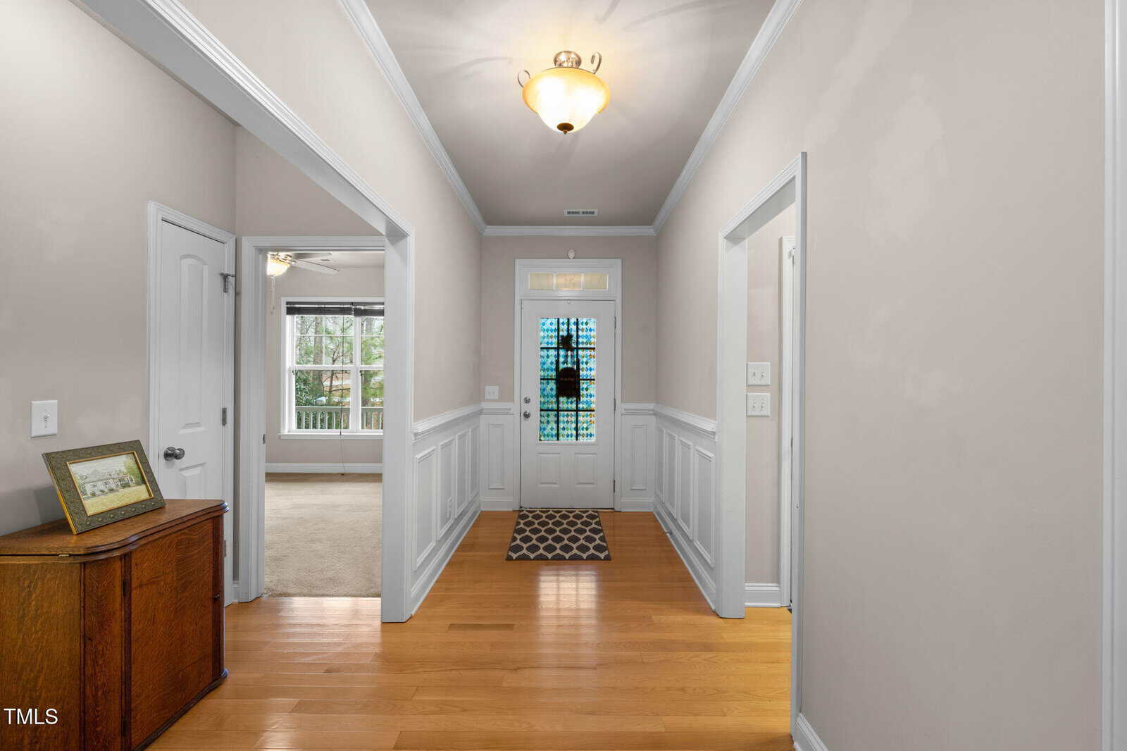 A bright hallway with light hardwood floors, beige walls, white trim, a hanging ceiling light, and a front door with glass panels. A small chest with a photo frame sits on the left, and rooms are visible on both sides.