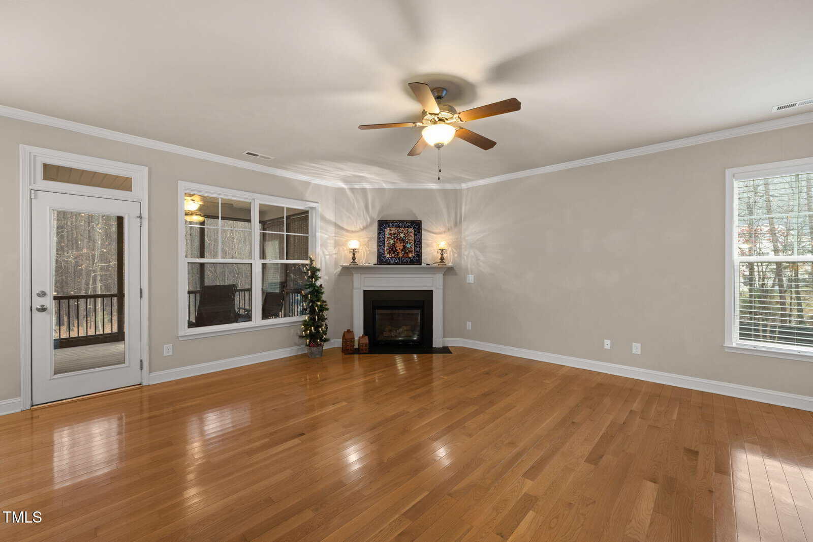 A spacious living room with hardwood floors, a ceiling fan, a fireplace with a mantel, two candles, and a decorative framed piece. Large windows and a glass door provide natural light and outdoor views.
