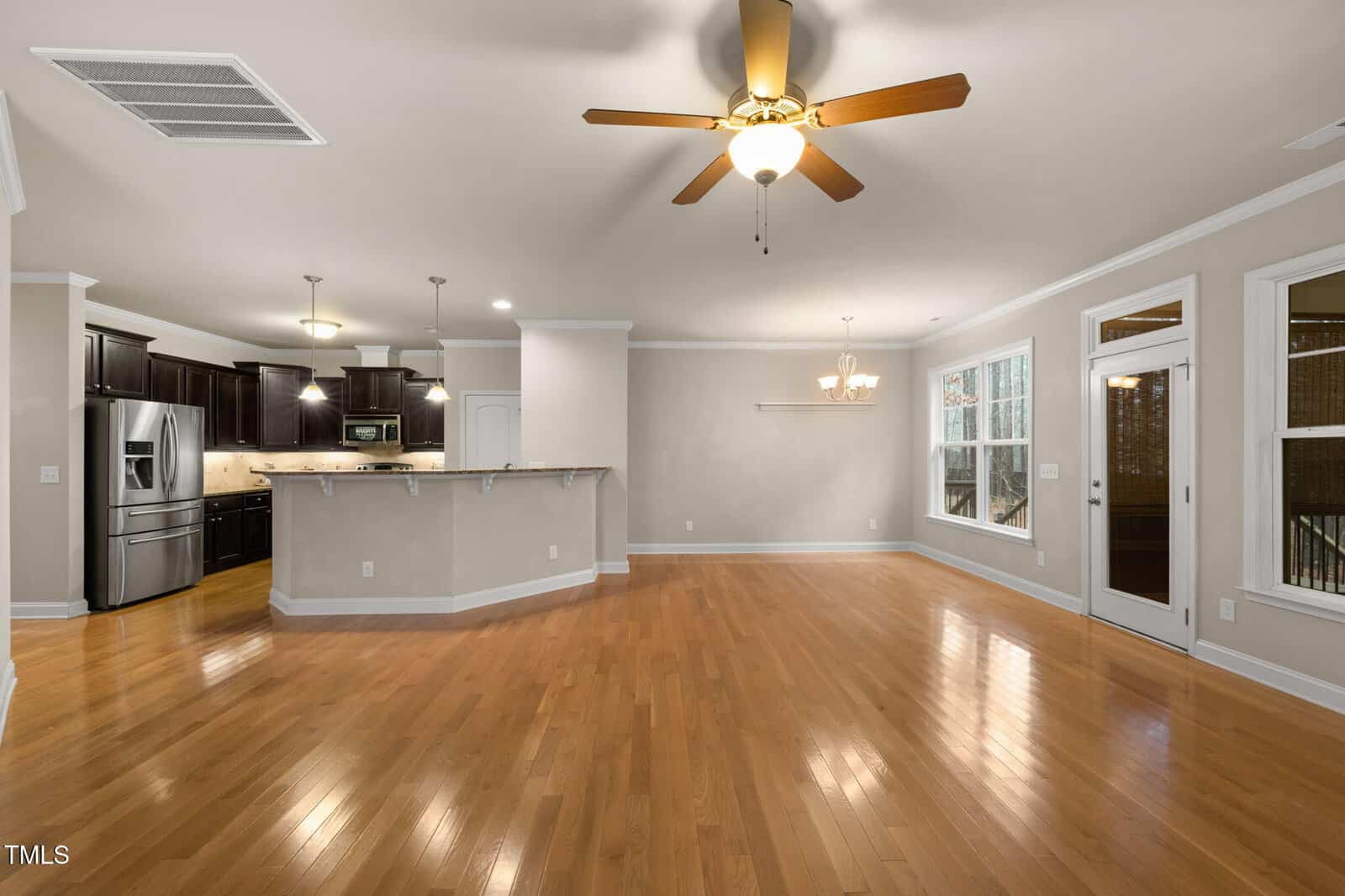 Open-concept living space with wood floors, ceiling fan, and large windows. The kitchen features dark cabinets, stainless steel appliances, and a breakfast bar with pendant lights. Dining area has built-in shelves.
