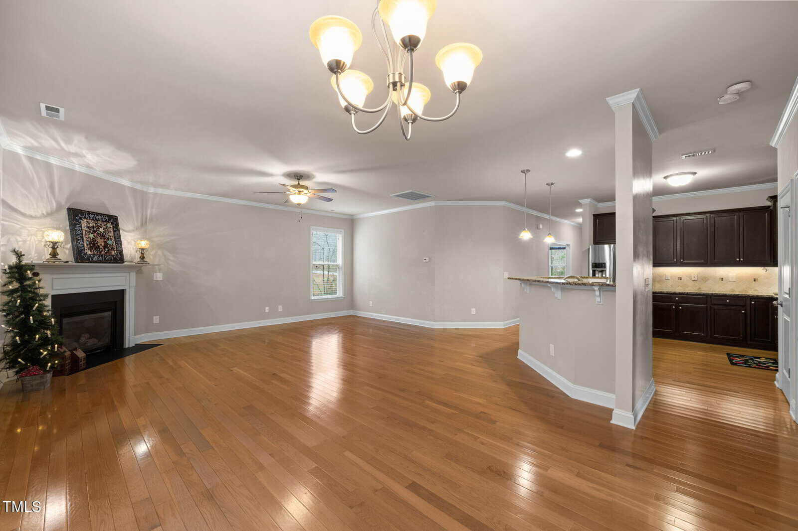 Spacious living area with hardwood floors, chandelier, fireplace, small Christmas tree, and open kitchen with dark cabinets and granite counters. Light gray walls and large windows brighten the room.