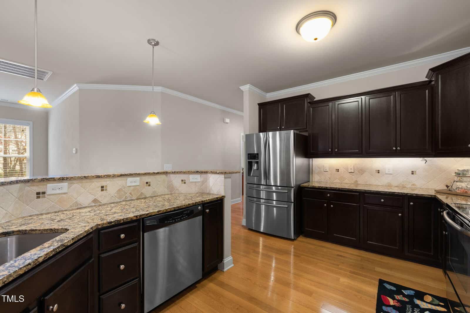 A modern kitchen with dark wood cabinets, stainless steel appliances including a refrigerator and dishwasher, granite countertops, tile backsplash, and hardwood floors under warm lighting.