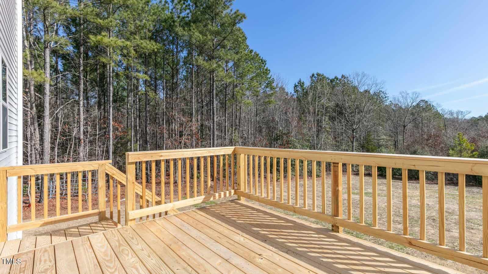 A wooden deck with railing overlooks a grassy backyard and a wooded area with tall trees under a clear blue sky.