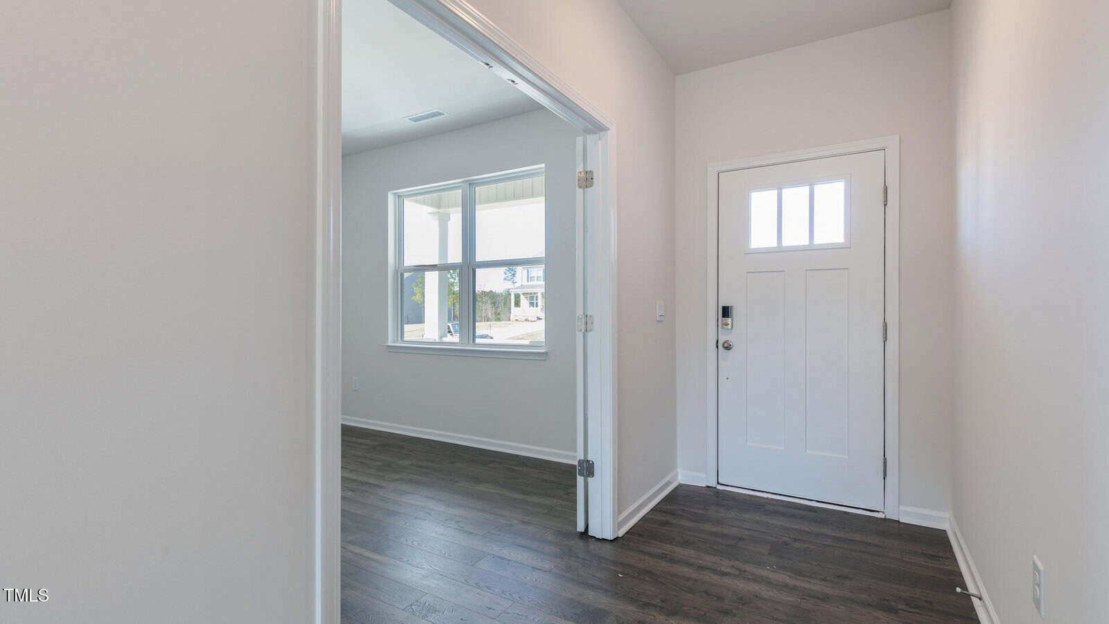 A white front door with a small window is beside a light switch and lock system. The entryway has dark wood flooring and light walls. An open doorway leads to a bright room with large windows.
