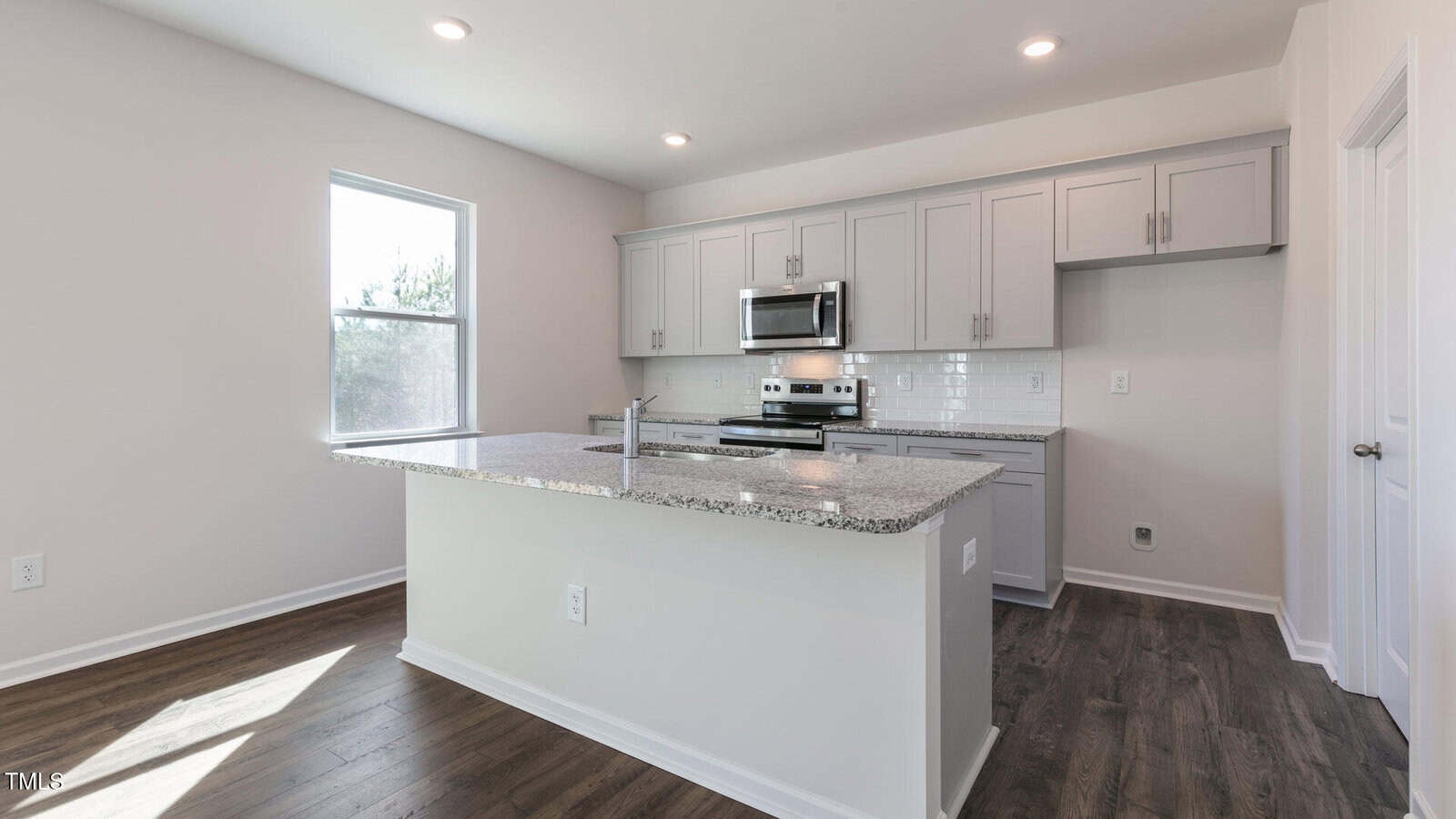 A modern kitchen with light gray cabinets, stainless steel appliances, white subway tile backsplash, granite countertops, and a center island with a built-in sink. The floor is dark wood, and there is natural light from a window.