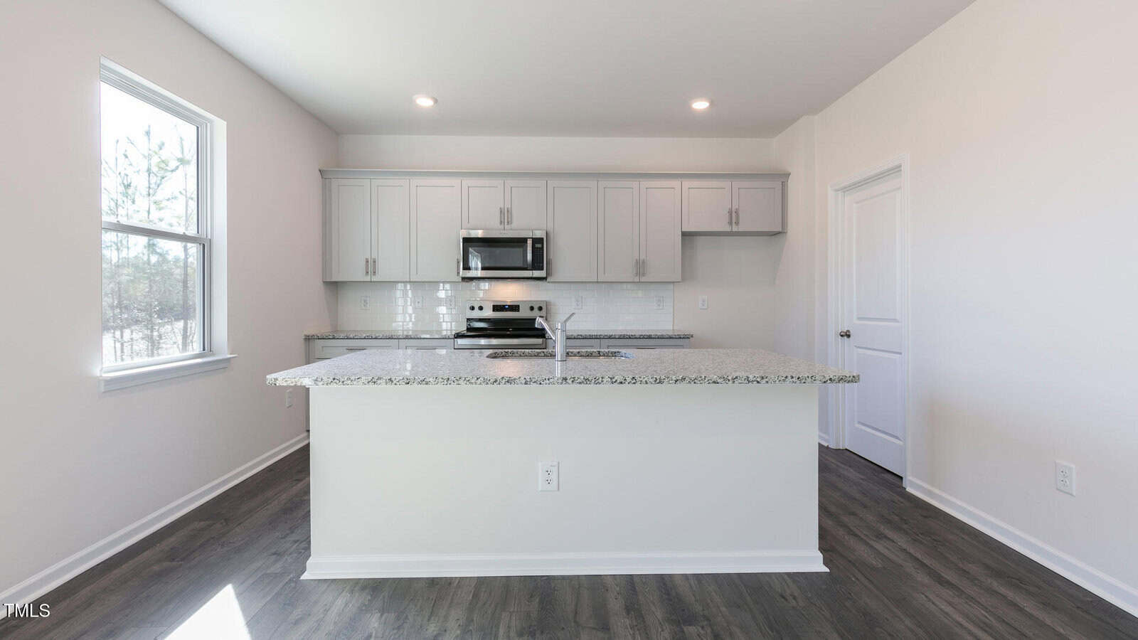 A modern kitchen with white walls, gray cabinets, stainless steel appliances, granite countertops, a large island, and dark wood floors. A window on the left lets in natural light.