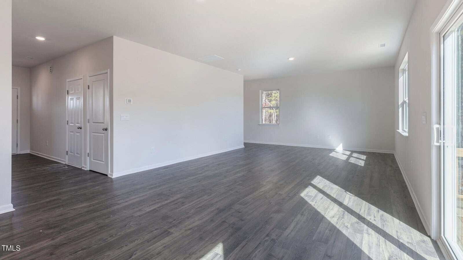 Bright, empty living space with gray wood floors, white walls, and large windows letting in natural light. Three doors and a hallway are visible on the left, and a glass door leads outside on the right.