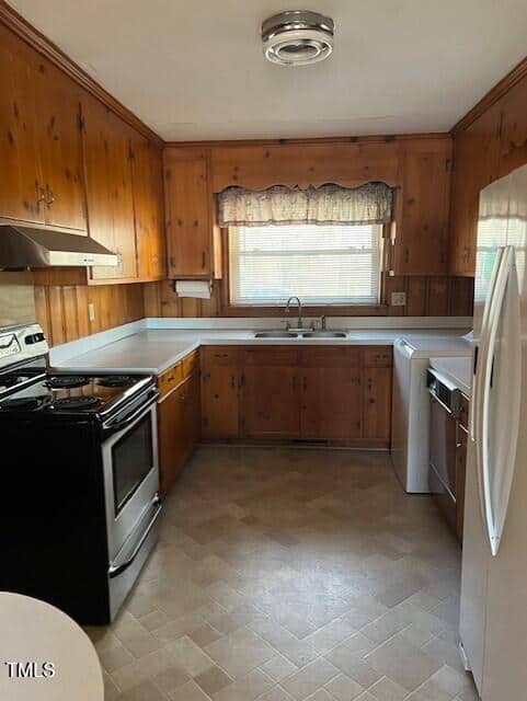 A kitchen with wood-paneled walls and cabinets, a stainless steel stove, dishwasher, white refrigerator, and washing machine. There is a double sink under a window with a valance, and beige tile flooring.