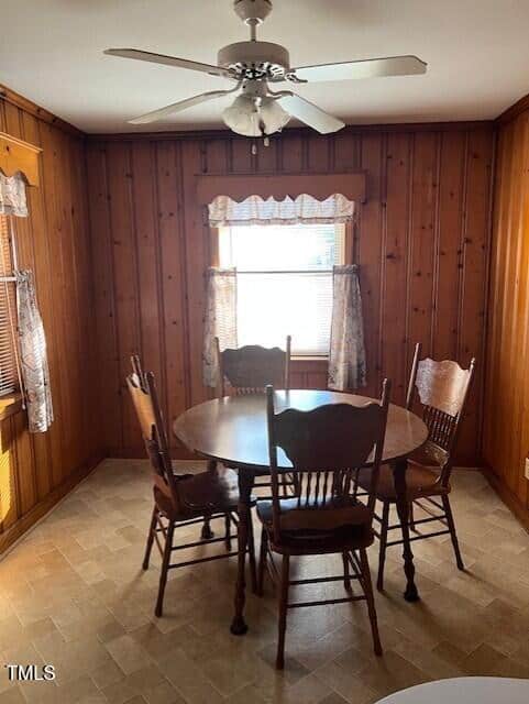 A small dining area with wood-paneled walls, a round wooden table, and four matching chairs. Two windows with sheer curtains let in natural light. A ceiling fan with lights hangs above the table.