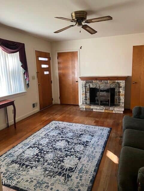 A living room with hardwood floors, a patterned area rug, a stone fireplace, two wooden doors, a ceiling fan, a window with vertical blinds and a valance, and part of a sofa and small table visible.