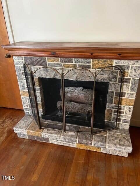 A stone fireplace with a wooden mantel and a black metal screen stands against a cream-colored wall on a hardwood floor. A wooden door is visible to the left.