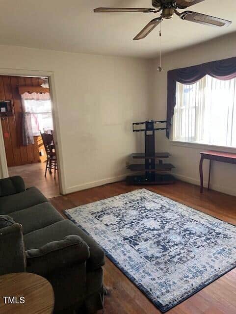 A living room with a couch, blue patterned rug, TV stand, side table, and a window with curtains. A ceiling fan hangs above, and a doorway leads to a dining area with wood-paneled walls.