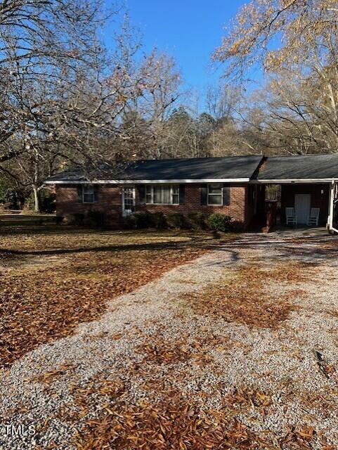 A one-story brick house with a covered carport, surrounded by trees with bare branches. The yard has scattered fallen leaves and a gravel driveway under a clear blue sky.