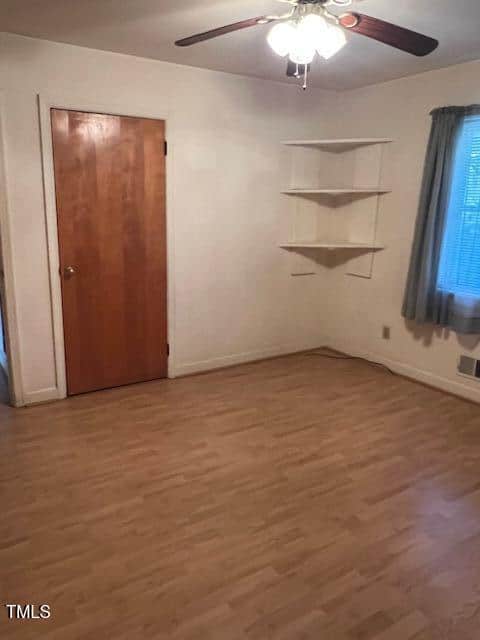 Empty room with light wood flooring, a ceiling fan with lights, a wooden door, a window with curtains, and white corner shelves on the wall. The walls are painted light beige.
