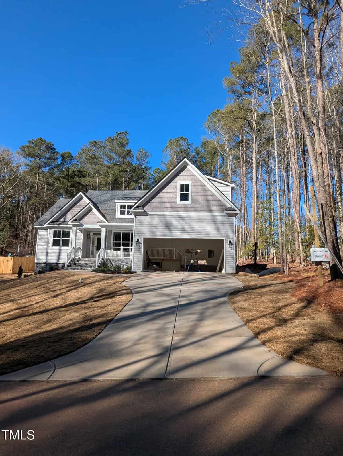 A gray two-story house with a double garage, long driveway, and small front porch is surrounded by tall trees and a clear blue sky.