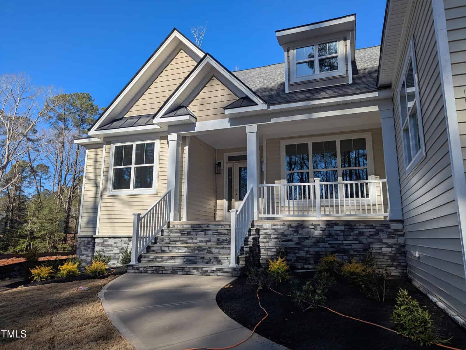 A modern beige house with white trim, large windows, stone steps, and a covered front porch with railings. Small shrubs line the walkway, and the sky is clear and blue.