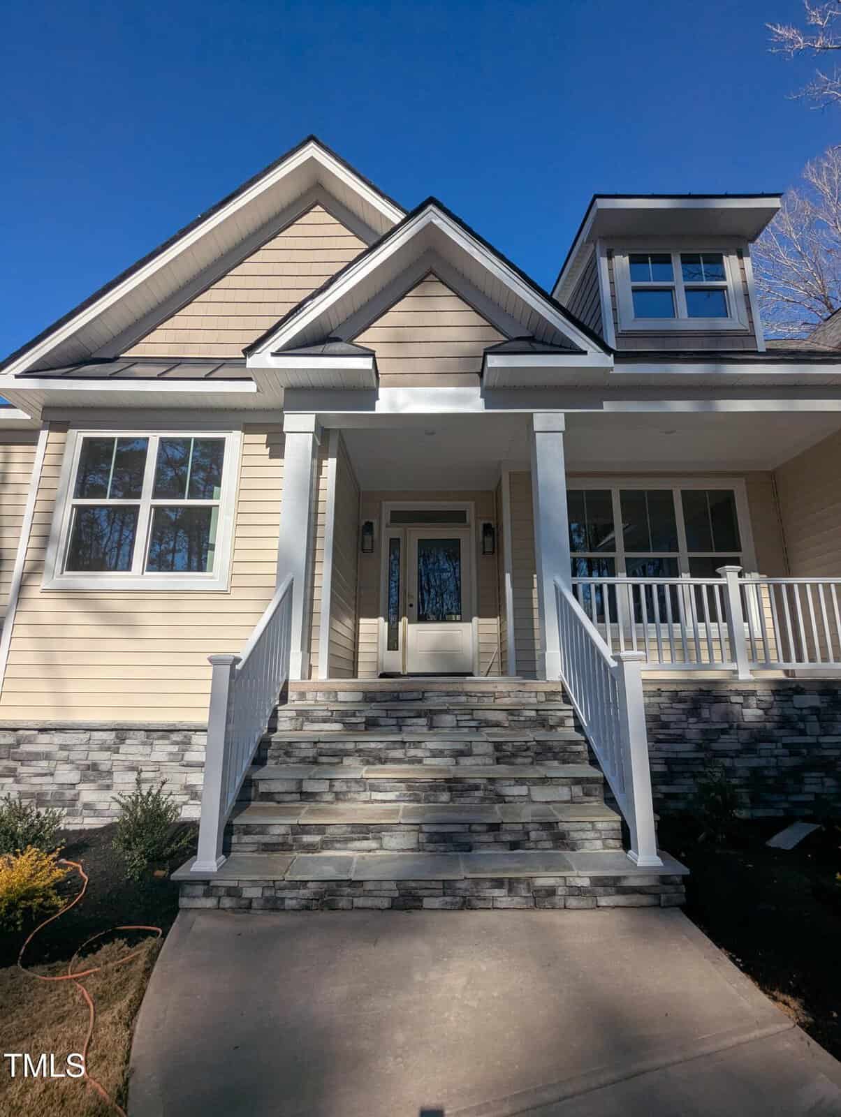 Front view of a modern beige house with stone steps, white railings, a covered porch, large windows, and a gable roof, set against a clear blue sky.