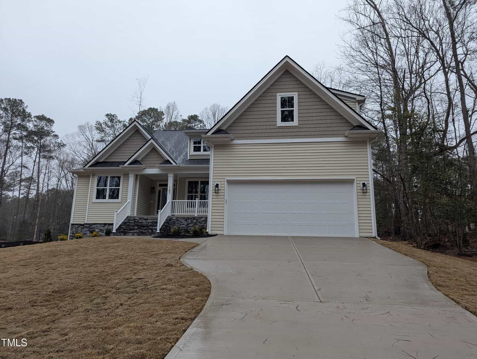 A two-story beige house with white trim, a front porch, and a large two-car garage sits at the end of a sloped concrete driveway, surrounded by brown grass and trees on a cloudy day.