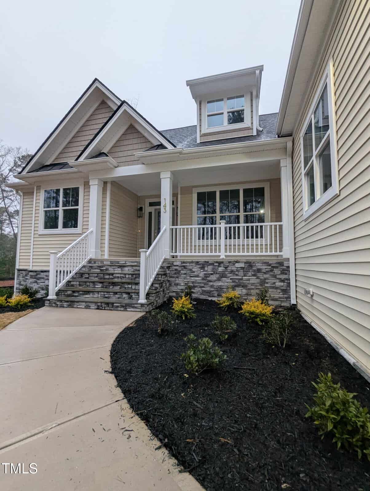A beige house with white trim, a covered front porch with white railings, gray stone accents, and steps leading to the entrance. There is a landscaped area with mulch and small shrubs next to the walkway.