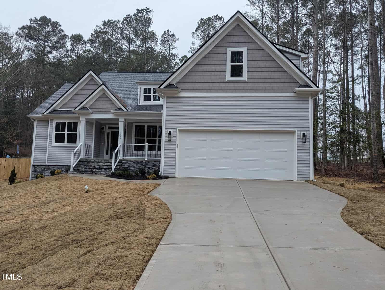 A two-story suburban house with gray siding, white trim, and a double garage. The front yard has straw-covered grass, a concrete driveway, and a covered front porch with steps. Pines trees are in the background.