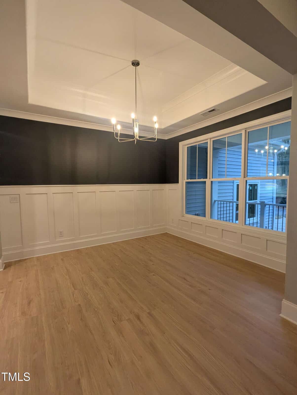 A modern, empty dining room with light wood floors, white wainscoting, a black accent wall, large window, and a contemporary ceiling light fixture.