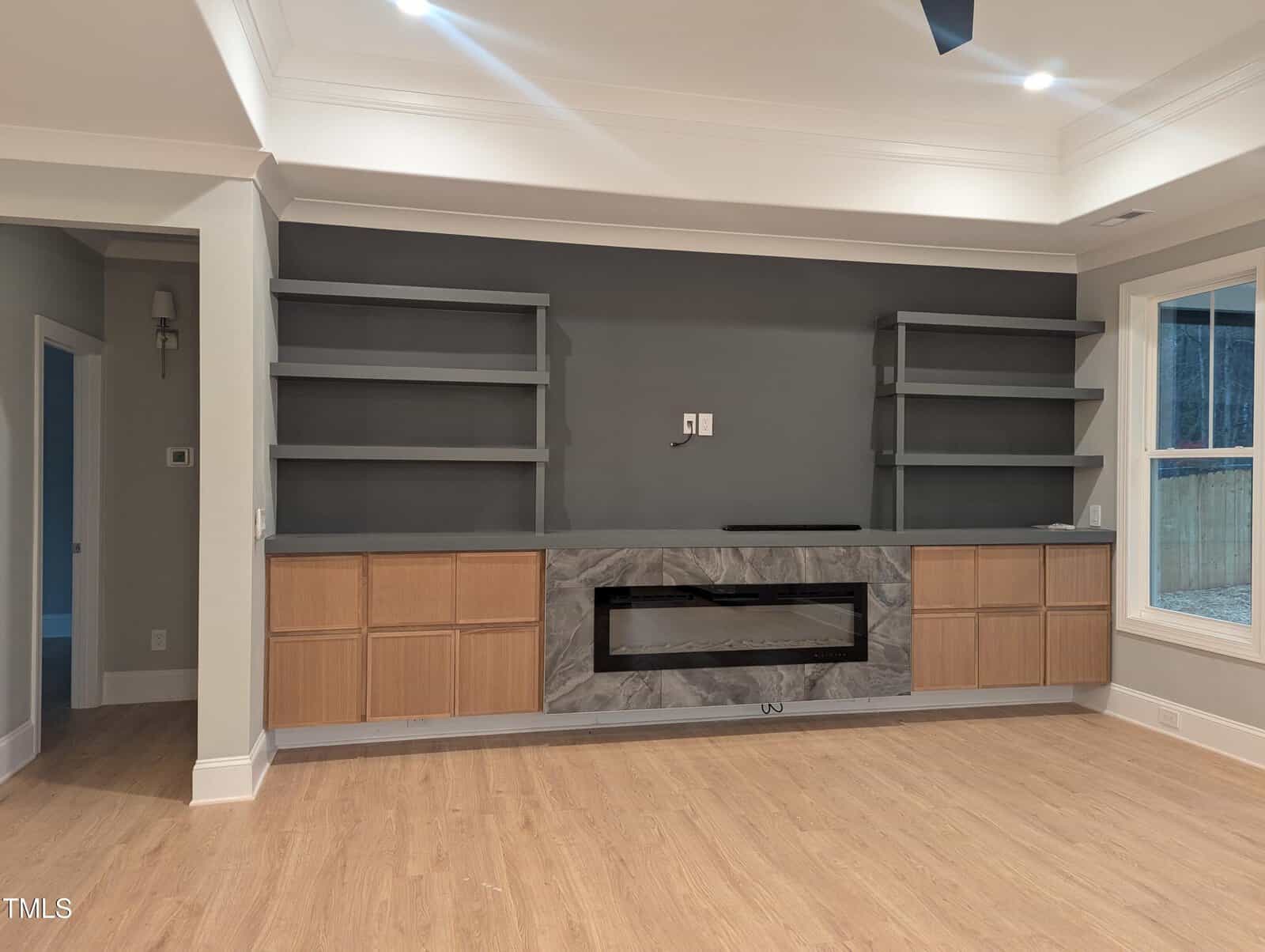Modern living room with built-in wooden cabinets, gray shelves, and a large electric fireplace set in a marble-patterned surround against a dark accent wall. Light hardwood floors and large window on the right.