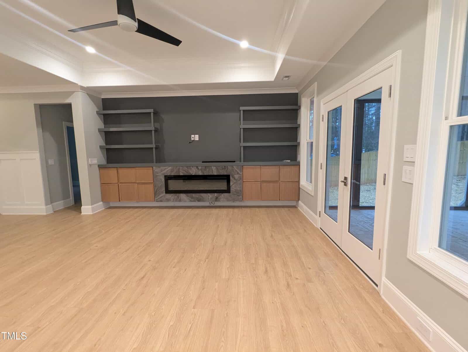 Modern living room with light wood floors, gray walls, a ceiling fan, built-in shelves, a fireplace, and glass doors leading to a patio. The room is bright with large windows and recessed lighting.