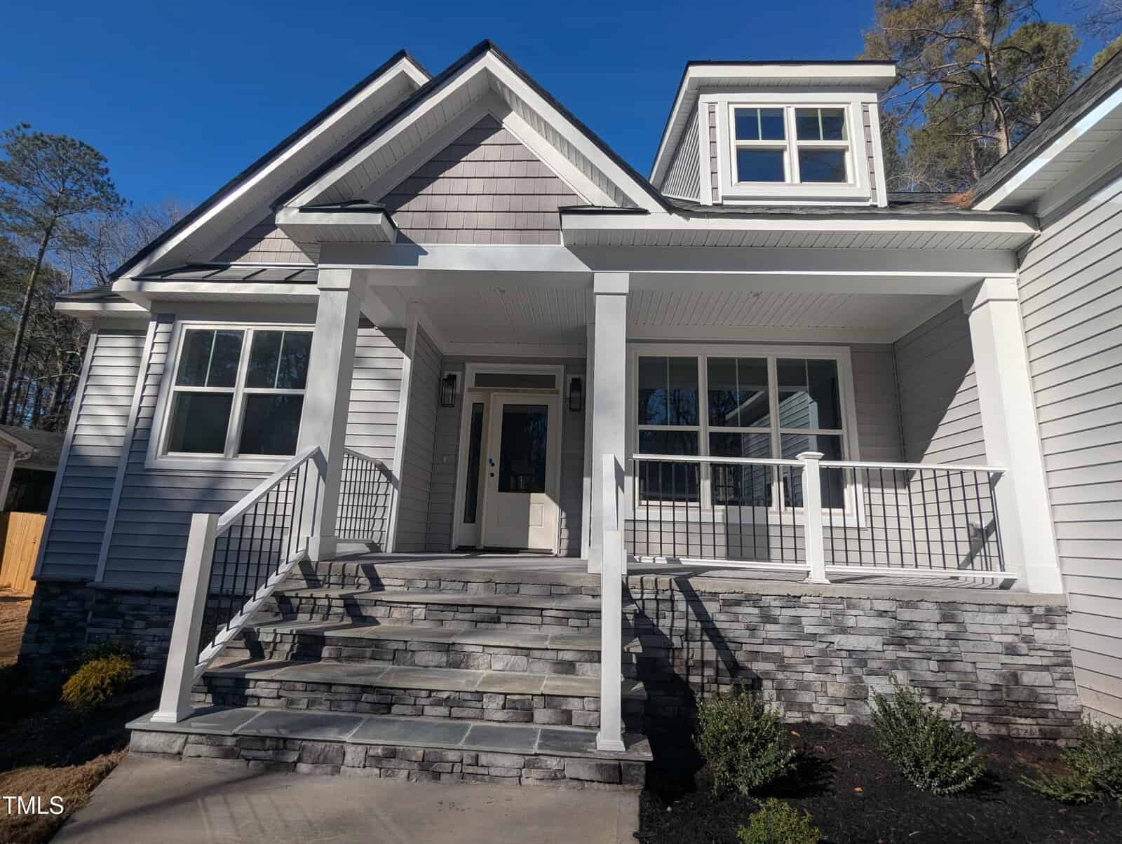 A front view of a modern house with light gray siding, multiple windows, a white front door, a covered porch with white railings, and stone steps leading up to the entrance. Shrubs line the walkway.
