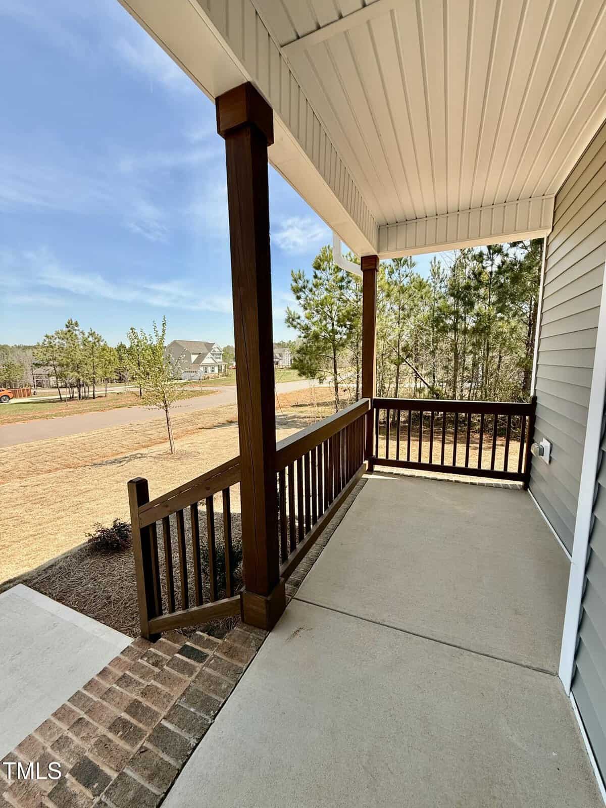 A covered front porch with wooden railings and columns, concrete flooring, and a view of a grassy yard, trees, and a suburban street under a clear sky.