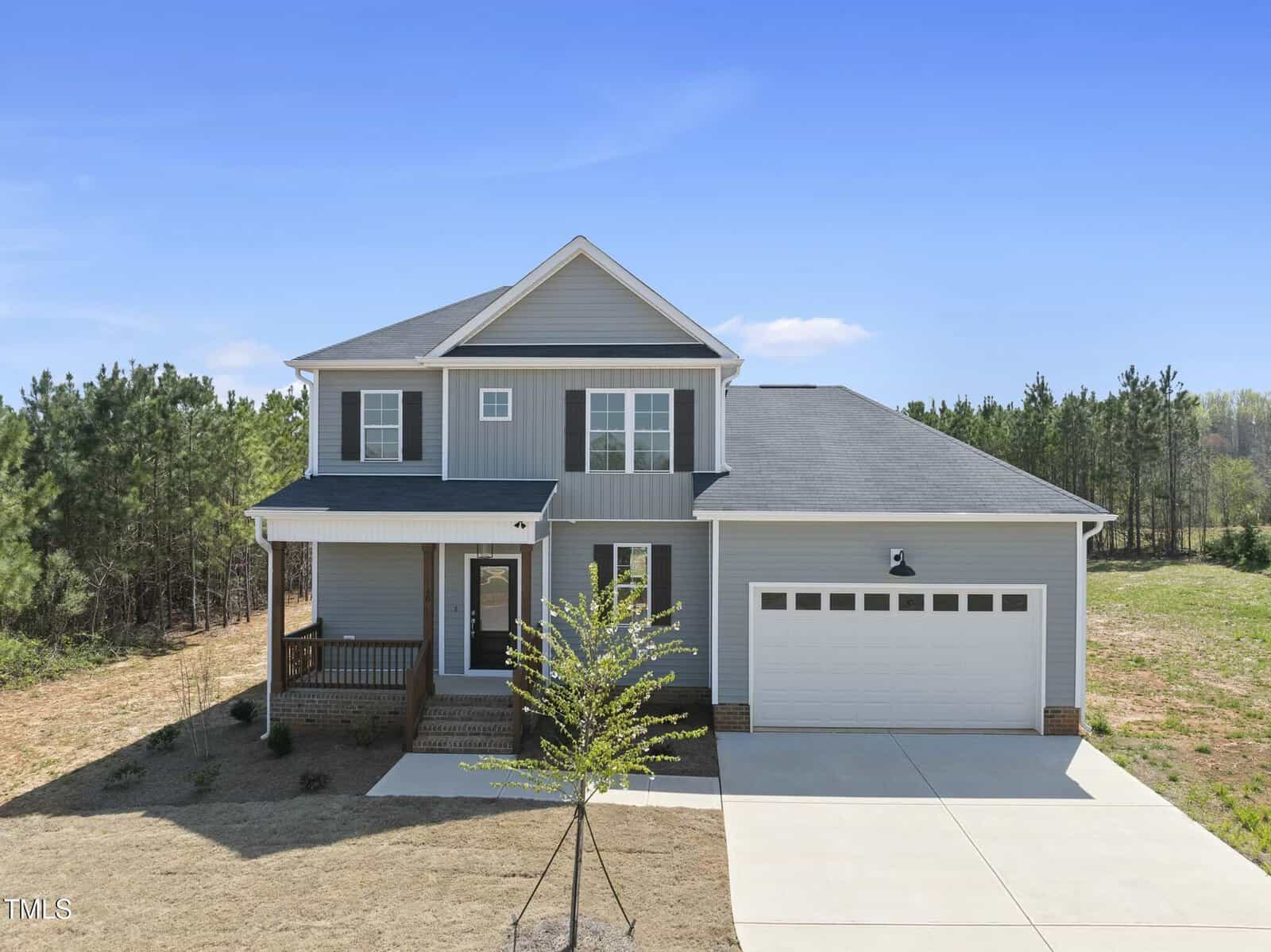 A two-story, gray house with white trim, a covered front porch, a two-car garage, and a small tree in the front yard. The house is set against a backdrop of pine trees and a clear blue sky.
