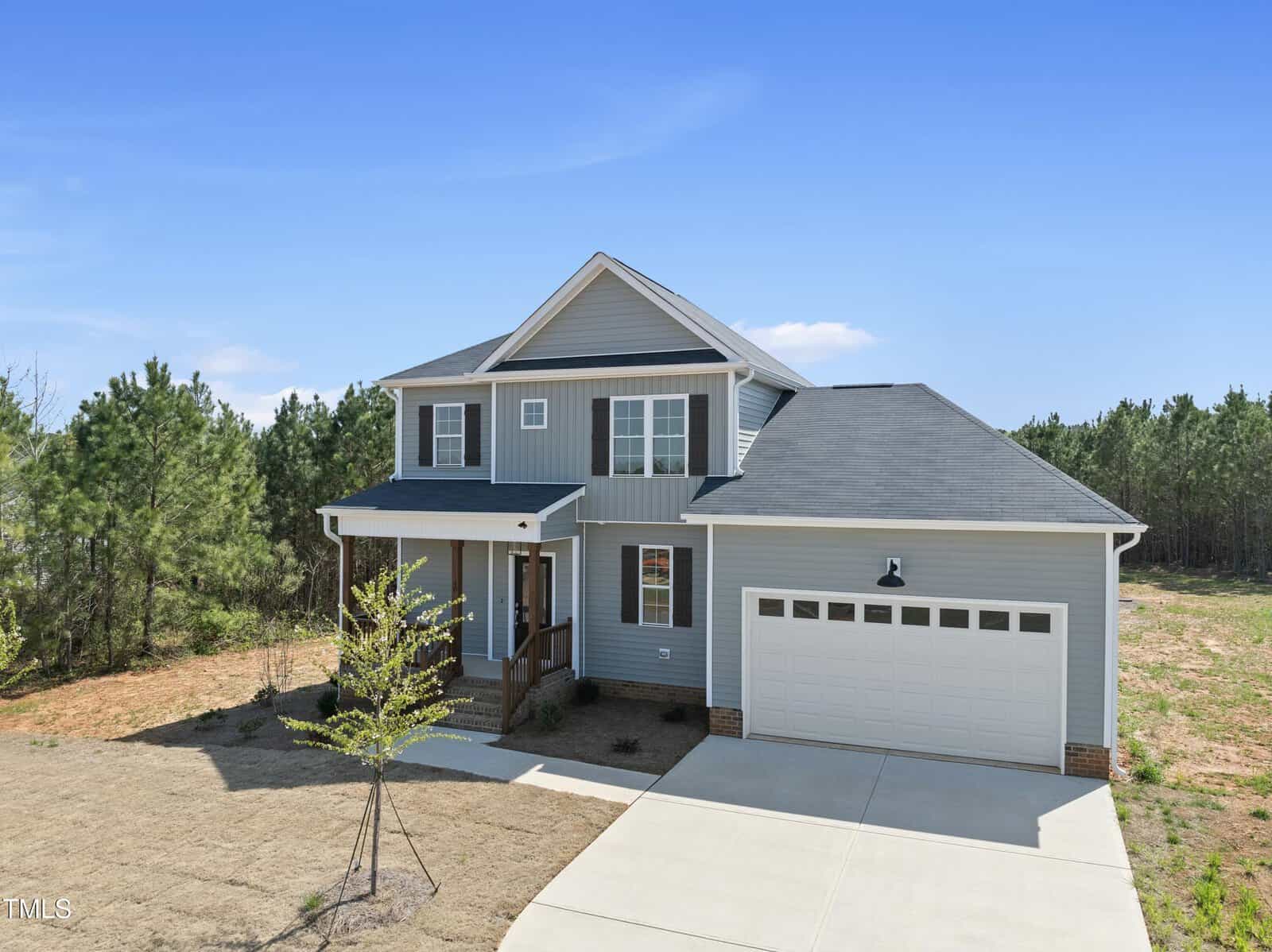 A two-story light gray house with white trim and a covered front porch, featuring a double garage and a concrete driveway, is situated on a mostly bare lot with a few young trees and surrounded by pine trees.