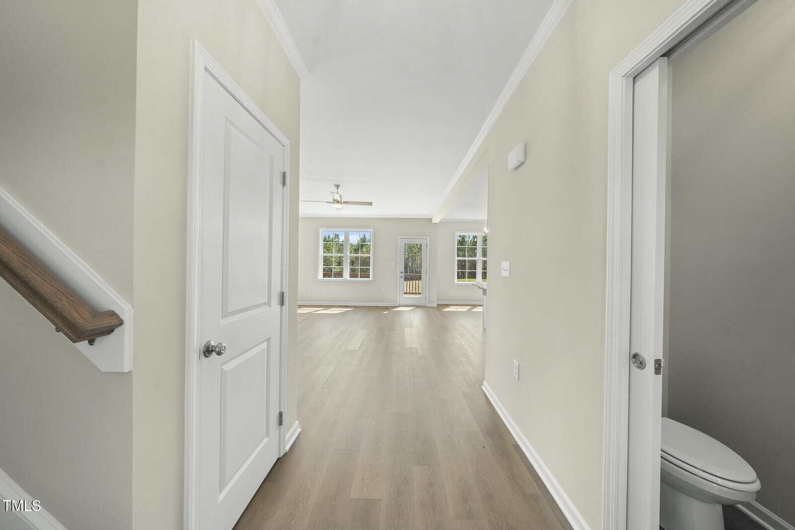 A hallway with light wood flooring leads to an open living area with large windows and a ceiling fan. There is a staircase on the left and a small bathroom with a toilet on the right.