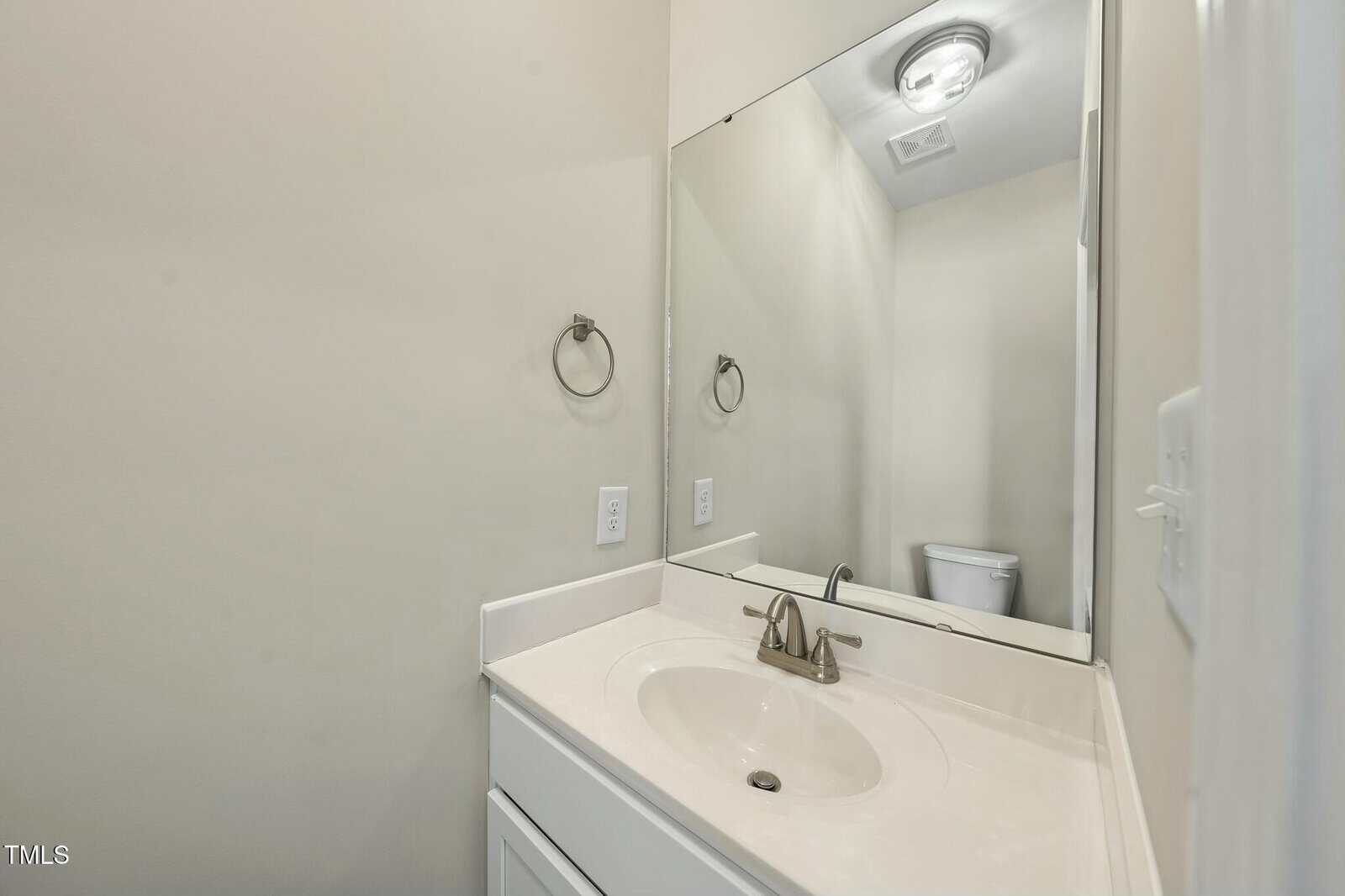 A small bathroom with a white sink vanity, silver faucet, and a large mirror above. There is a towel ring on the wall and a toilet reflected in the mirror. The walls are light beige.