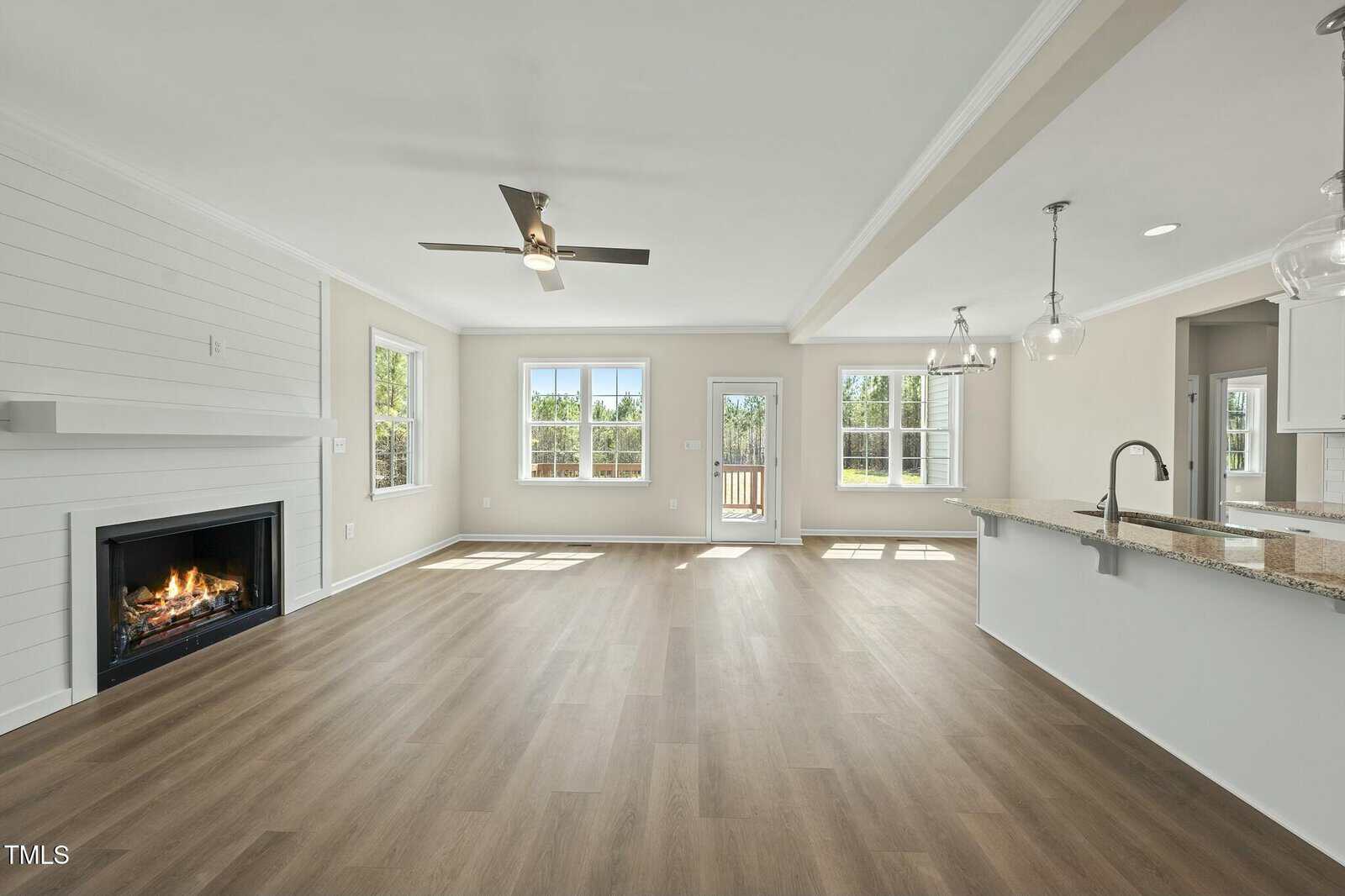 Bright open living space with large windows, a ceiling fan, a fireplace, light wood floors, and an adjoining modern kitchen with granite countertops and pendant lights. Door opens to a patio outside.