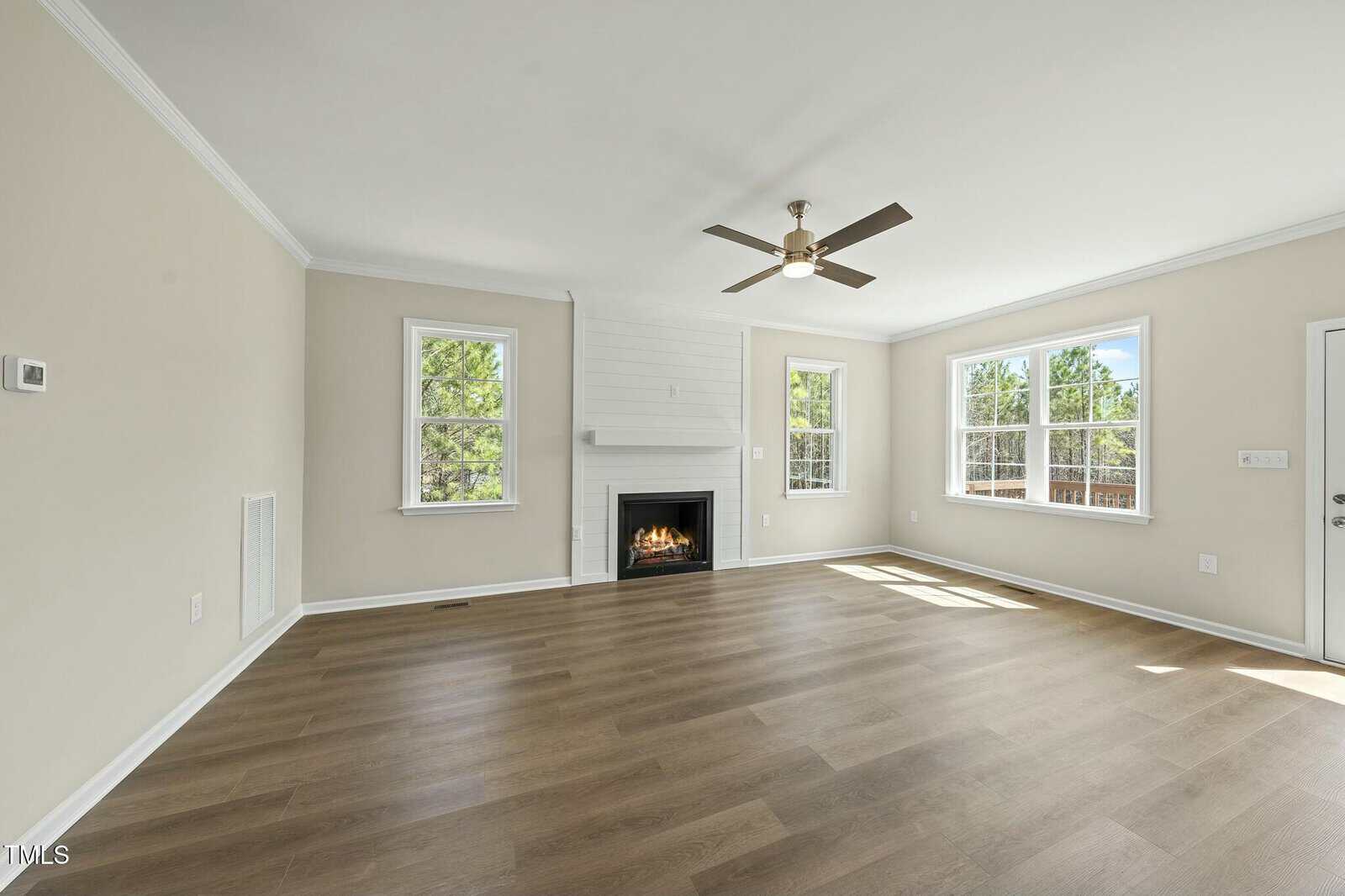 Bright living room with light wood flooring, beige walls, a ceiling fan, corner fireplace, and multiple large windows letting in natural light.