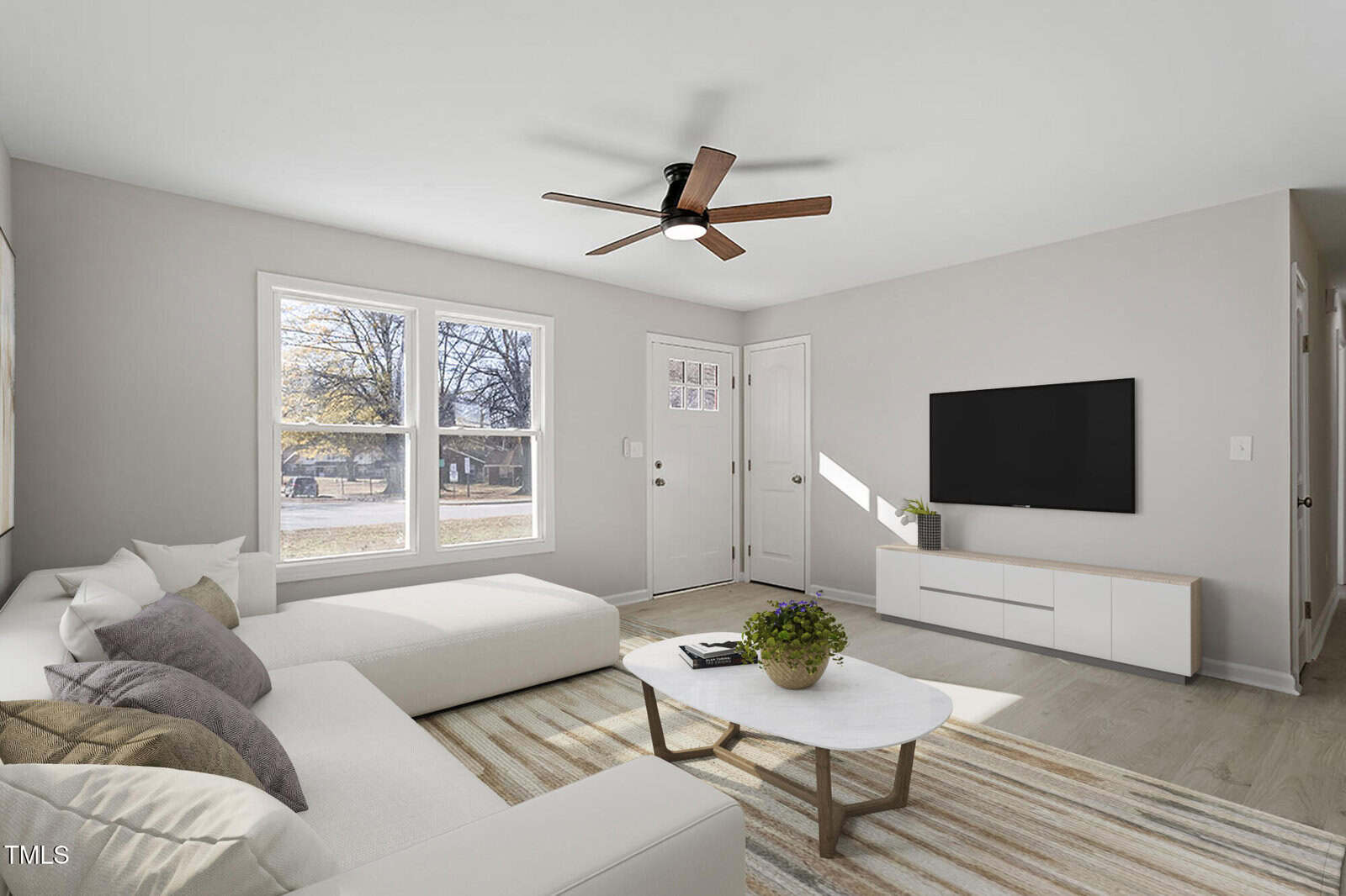 A modern living room with a white sectional sofa, gray and purple cushions, a round coffee table with a plant, wall-mounted TV, and a beige striped rug. The room features a ceiling fan and large windows with a view of a tree-lined street.