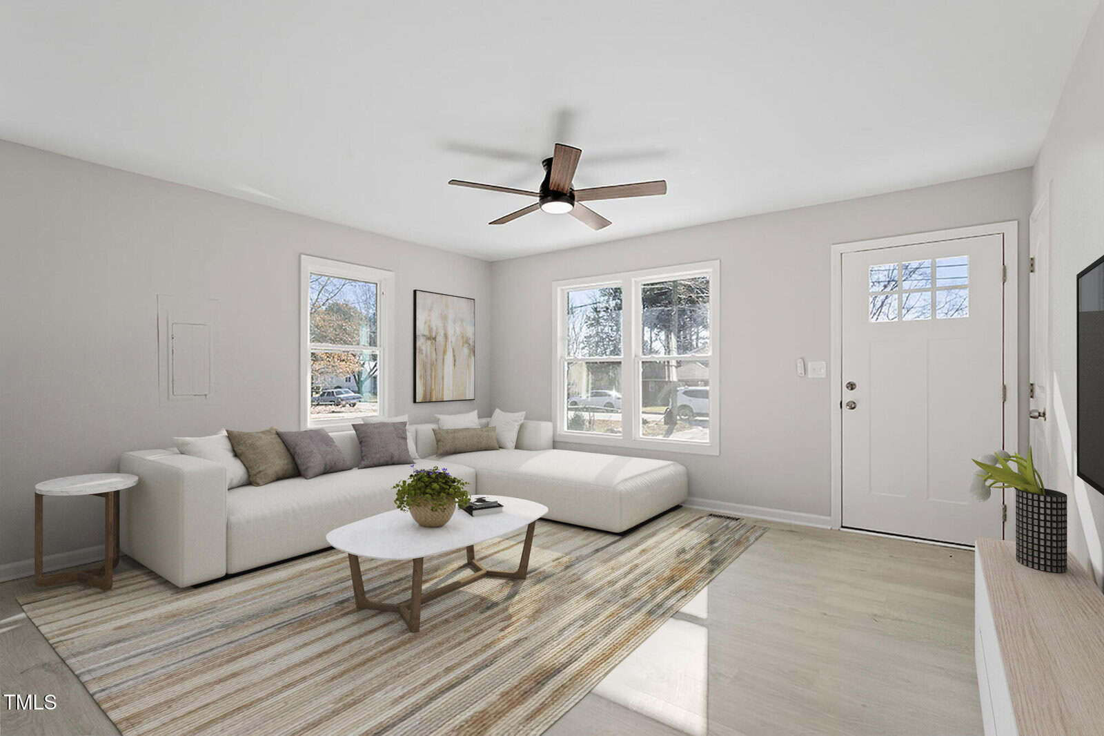 A modern living room with white walls and light wood flooring. It features a large L-shaped sofa, a round coffee table with plants, a ceiling fan, and a rug. Three windows overlook a wooded area, and a door is next to the windows. A wall-mounted TV is on the right.