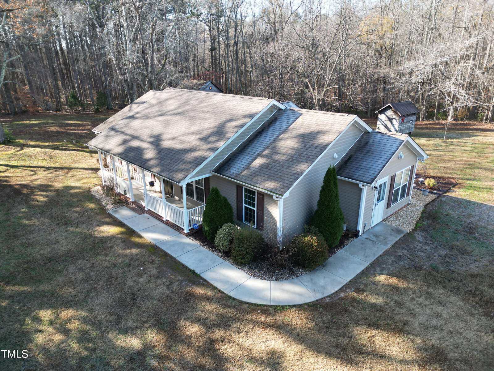 An aerial view of a single-story house with a gabled roof, surrounded by a manicured lawn and trees. A sidewalk leads to the front entrance, and a small structure is visible in the background amidst wooded surroundings.