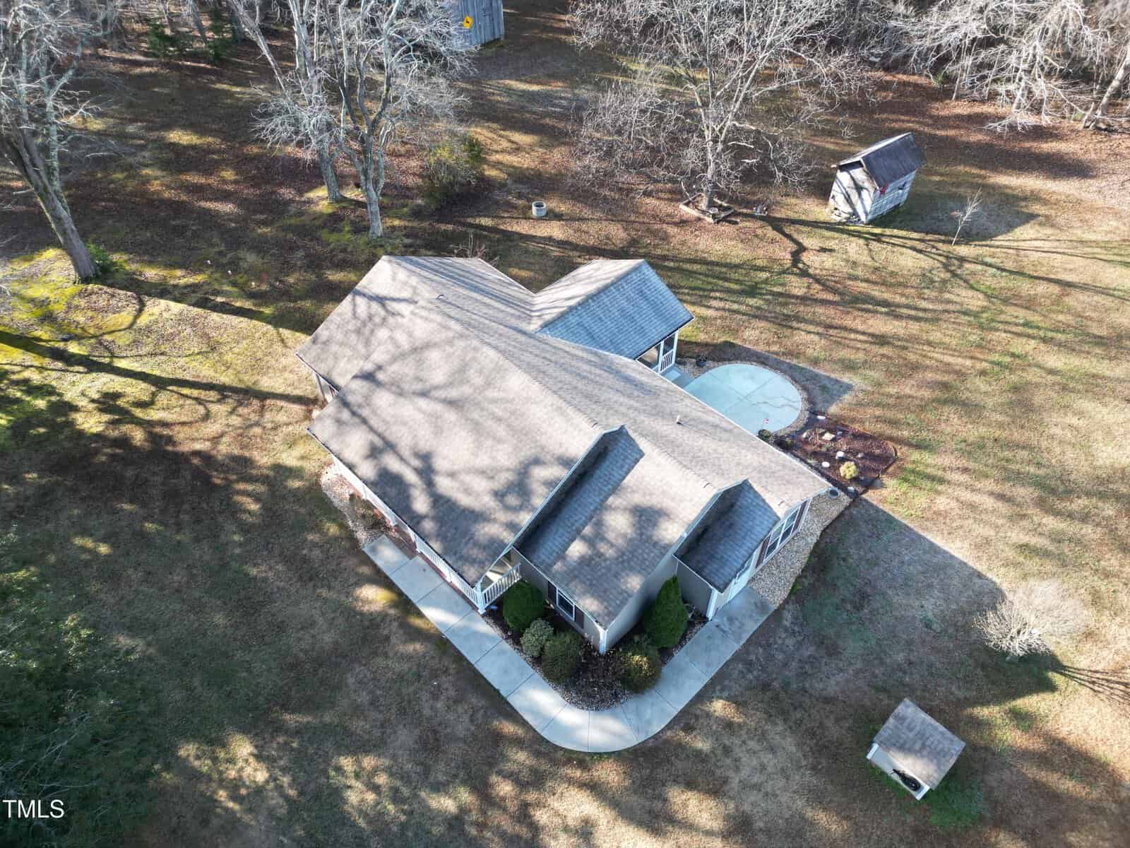 Aerial view of a single-story house with a gray roof, surrounded by a lawn and trees without leaves. A curved walkway leads to the front entrance. A small shed is visible in the backyard. Shadows of bare branches are cast on the ground.